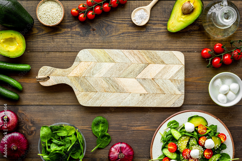 Wall mural Cooking fresh salad. Vegetables, greens, spices, plate of salad, cutting board on dark wooden kitchen desk top view mockup