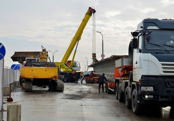 A crane unloads a long concrete slab from a truck at a construction site. Workers unload reinforced concrete panels