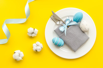 Fototapeta na wymiar Easter table decoration. Plate, cutlery, painted eggs and dry white flowers, tablecloth on yellow background top view