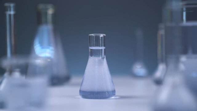 On the table is a medical flask with liquid. The camera smoothly leaves the flask. Gray wall on background. Flasks in defocus on the gray background.