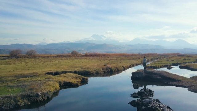 drone dolly forwards, bypassing a traveller standing at edge of rock, looking at scenic view, next to pond, cloudy weather, mountains in the background, in Kayseri, Turkey