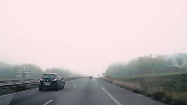 Dashboard camera view driving on a French highway as cars pass green trees and graffiti covered barrier by on a foggy misty day in slow motion