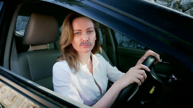 a portrait of an androgynous feminist with a painted mustache on her face sitting behind the wheel of a car. woman looks into the camera from the car window.