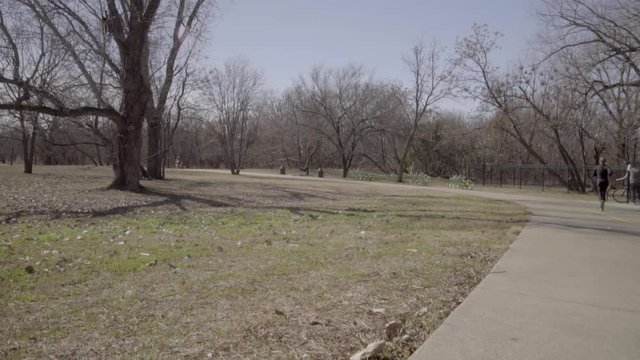 This is a wide shot of a jogger and a cyclist exercising in a park. The jogger and cyclist pass the camera while a Dog and his Owner are seen in the background.