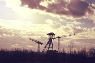 Silhouettes of old coal mine tower and two cranes against dramatic skyscape in Genk, Belgium. Industrial construction and mining concept.