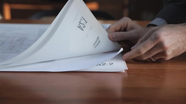 Architect flipping through a set of drawings on a wooden table in natural daylight. Close up.