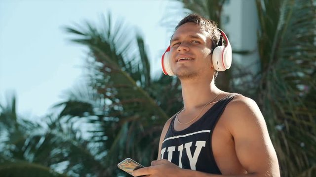 Handsome Young Man Listening Music from his Smartphone in Wireless White Headphones, Dancing on Beach Urban at Background Blue Sky.