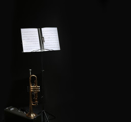Trumpet, case and note stand with music sheets on black background. Space for text