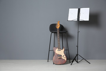 Electric guitar, chair and note stand with music sheets near grey wall indoors. Space for text