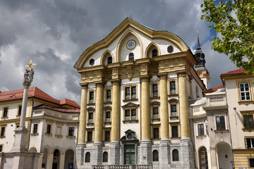 Fototapeta premium Full sun on the Ursuline Church of the Holy Trinity with marble statues of the Holy Trinity column in Ljubljana Slovenia with dark clouds