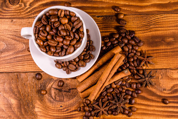 White cup filled with coffee beans, star anise and cinnamon sticks on wooden table. Top view
