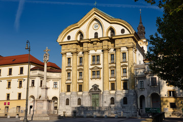 Fototapeta na wymiar Sunshine on the Ursuline Church of the Holy Trinity with marble statues of the Holy Trinity column in Ljubljana Slovenia with clock and bell tower
