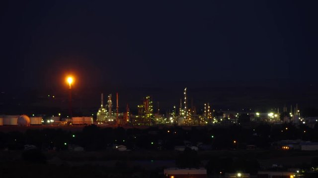 Twilight falls on an oil refinery with flaming tower and lights.