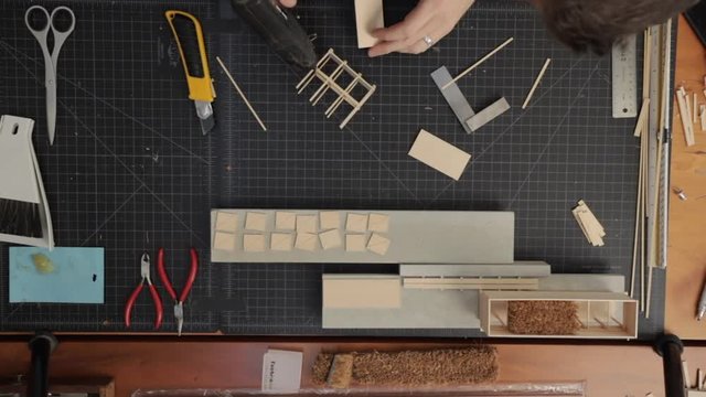 Overhead shot of a male architect gluing and constructing a scale model of a house in his design studio. Background is a black gridded cutting mat.
