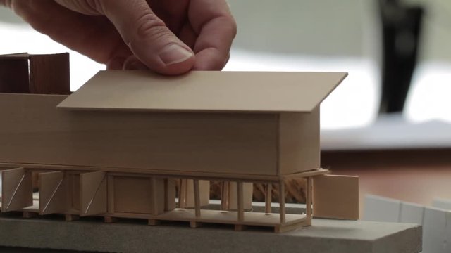 Close up view of male architect's hands testing out a roof shape on a wooden scale model of a modern home design. Backlit shot.