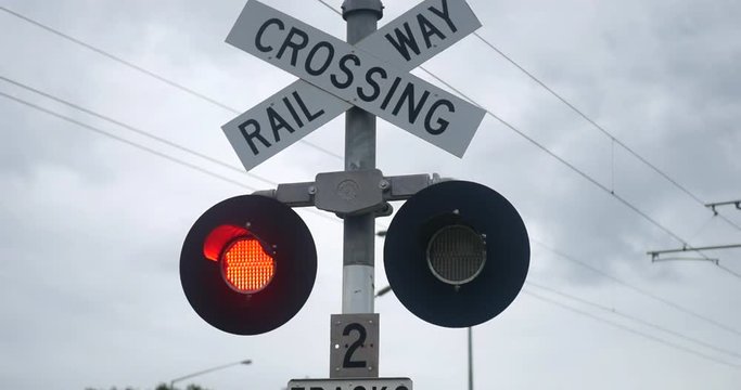 Sliding shot of a rail way crossing sign with two red lights flashing and trees in the background, in a cloudy day