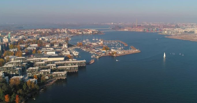Aerial, reverse, drone shot, above the sea, of buildings and boats, at Lauttasaari harbor, on a sunny, fall day, in Helsinki city, Finland