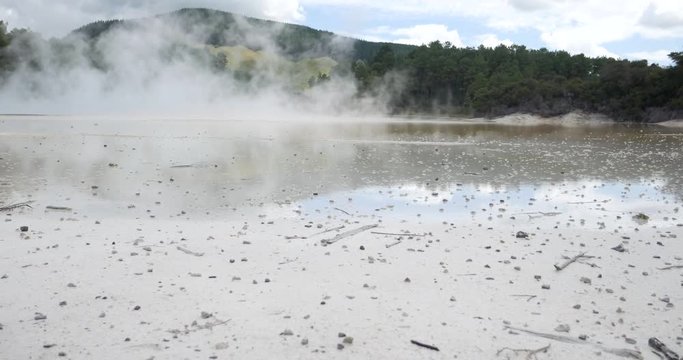 Sliding shot of Wai-O-Tapu Thermal Wonderland, New Zealand, with steam going up from the water and trees and mountains in the background, on a sunny day with some clouds