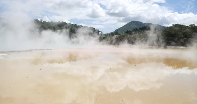 Panoramic shot of Wai-O-Tapu Thermal Wonderland, New Zealand, with steam going up from the water and trees and mountains in the background, on a sunny day with some clouds
