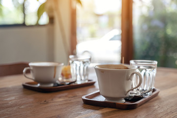 Closeup image of white mugs of hot coffee and glasses of water on wooden table in cafe