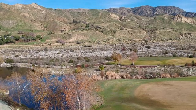 A Drone raises up from behind a bush to reveal mountains and 3 golf carts speeding along a path on a beautiful golf course.