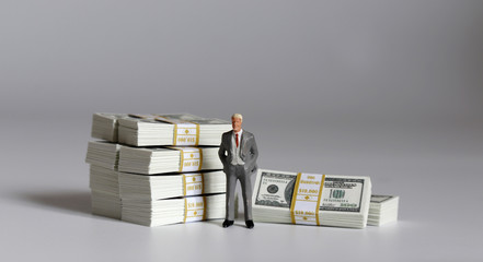 A miniature businessman standing with a pile of $100 bills.