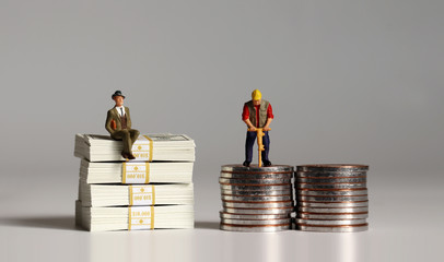 A miniature businessman sitting on a pile of $100 bills and a miniature worker on a pile of coins.