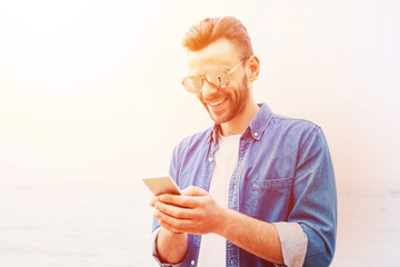 Sunshine stories. Happy man in denim shirt and white t-shirt is using his mobile phone on the sea shore in front of marvelous sun set.