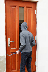 Robber in hood trying open door with crowbar and looking through glass pane, back view. Crime, burglary and house robbery concept.