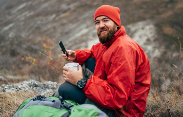 Young explorer bearded man drinking tea or coffee in mountains. Traveler man wearing red clothes...
