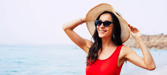 All charm and sunshine. Chic girl in front of light blue sea water is wearing bright swimsuit, taupe round hat and cat-shaped black sunglasses and welcoming the morning sun.