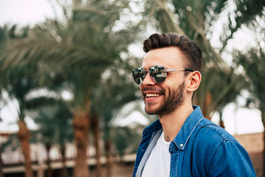 Catch The Moment. Chilly Man Near The Huge Palms, In Jeans Shirt And Sunglasses Is Happily Looking Away And Smiling Stunningly. 