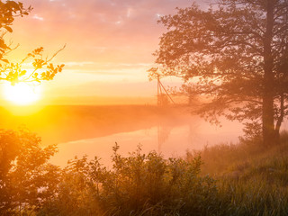 Fototapeta na wymiar Misty morning at the riverbank in summer. Rising sun shining though the fog