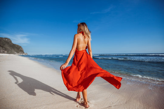 The girl is on the white sand on the beach in a red flying dress. Photo without Face, back to us.