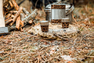 flask with glasses, camping woodstove and utensils, axe and sapper shovel near a firewoods, backpack on the background