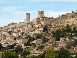 Typical Catalan village Guimera on a hillside. Light brown rectangular blocks of ancient stone buildings against a beige cliff, green trees and blue sky © ioanna_alexa