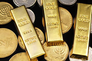 A macro image of miscellaneous coins from Israel with three small gold ingots on a reflective black background