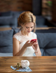 The bride with smartphone is sitting near  table with coins and dollars in moneybox