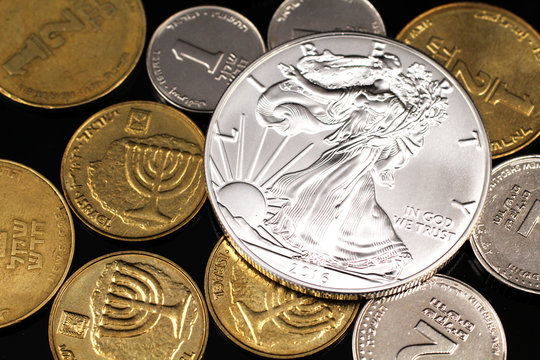 A macro image of miscellaneous coins from Israel with an American one ounce silver coin on a reflective black background