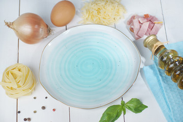 Empty blue plate on a white wooden table with ingredients spread over the edge for making carbonara paste.