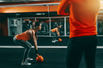 Young Caucasian sporty woman in orange shirt and ponytail lifting kettlebell in gym at night while her personal trainer looking at her. In background mirror. Excuses don't burn calories.