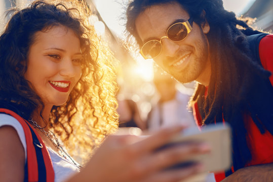 Cute smiling young woman with curly hair and backpack on backs showing to her mixed race friend something on smart phone. Summer vacations concept.