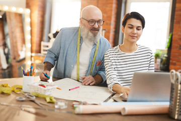 Obraz premium Two casual creative designers using laptop to search for new fashion trends in the net while working in studio