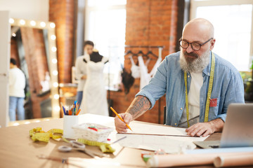 Senior tailor or fashion designer making sketch of new items for seasonal collection by workplace