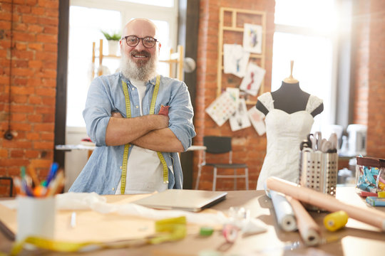 Cross-armed senior tailor in casualwear and eyeglasses standing by desk in front of camera in workshop