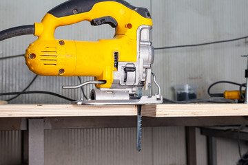 A yellow electric jigsaw ready for use stands in a sawn wood building material on a workbench in a workshop for carpentry work. Tool for industry and production.