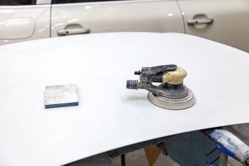 Grinding machine and emery sponge in the process of working on a car part of the hood of white color during preparation for painting after an accident in a body repair shop for vehicles