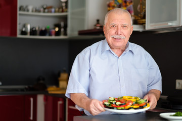 Smiling Senior man 70-75 yers old, holding plate with fresh vegetables on her kitchen at home.