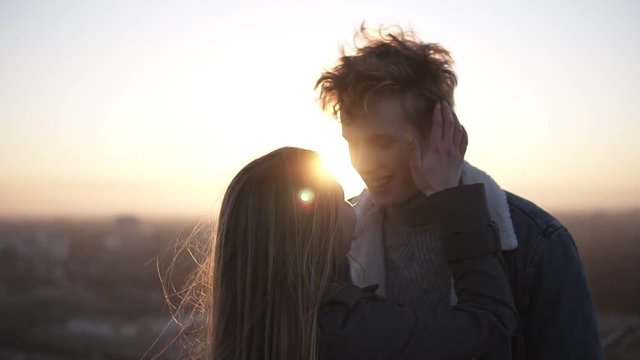 Outdoor close up portrait of young happy stylish couple hugging on the roof at sunset or sunrise. Girl caressing her boyfriend. Standing in front the the sunlight