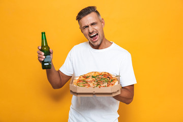Image of excited man 30s in white t-shirt drinking beer and eating pizza while standing isolated